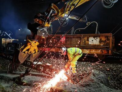 Mobix werker met gele hi-vis kleding aan het slijpen op het spoor bij nacht, met opspattende vonken